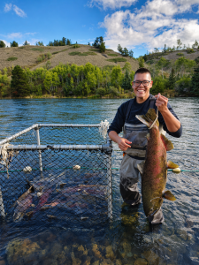 A person stands proudly in a river, holding a large fish near a metal enclosure containing more fish. The background features a hillside with lush green trees.